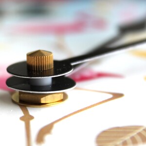 A close-up view of a metallic object with a golden-colored top, resting on a white surface with a pink and gold pattern.