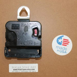 a black quartz clock with a silver hook, placed on a brown surface next to a circular sticker that reads "made in USA".