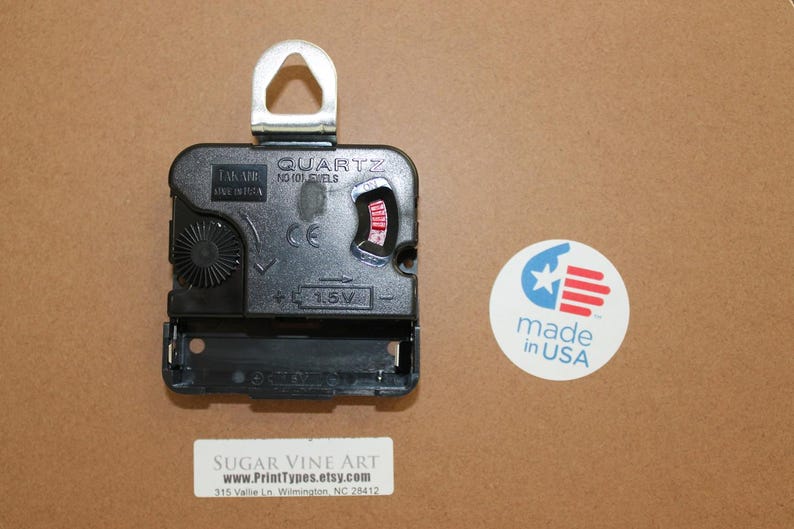 a black quartz clock with a silver hook, placed on a brown surface next to a circular sticker that reads "made in USA".