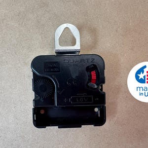 a black electronic component, likely a clock mechanism, with a silver hook attached to it. There is a logo or emblem next to the component, which appears to be a symbol of the United States.