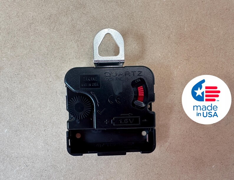 a black electronic component, likely a clock mechanism, with a silver hook attached to it. There is a logo or emblem next to the component, which appears to be a symbol of the United States.