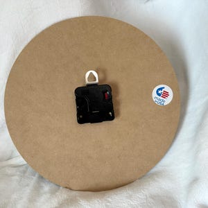 A round wooden clock with a black clock face and a white hook is placed on a white surface.