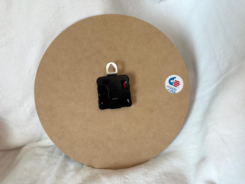 A round wooden clock with a black clock face and a white hook is placed on a white surface.