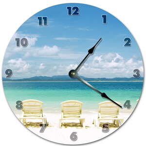 May include: A round clock with a beach scene. The clock face has white numbers on a blue sky background. The beach scene shows three white lounge chairs on a sandy beach with turquoise water and a blue sky with white clouds. The clock hands are black.