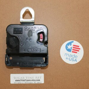 a black quartz clock with a silver hook, placed on a brown surface next to a circular sticker that reads "made in USA".