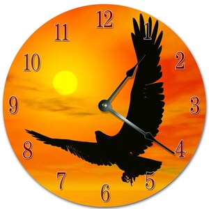 May include: Round wall clock featuring a silhouette of an eagle in flight against a sunset backdrop. The clock face has a gradient of orange and yellow, with red numerals marking the hours. The clock hands are silver.