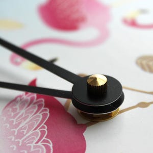 A close-up view of a clock face with a black dial and a gold-colored knob, set against a background with a pink flamingo and other abstract shapes.