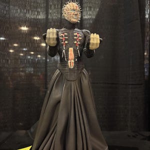 May include: A Pinhead action figure featuring a pale head covered in silver pins, wearing a black textured bodice with red accents and a long, flowing black skirt. The figure has both hands raised with clenched fists and a detailed torso design.