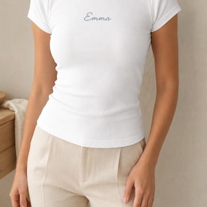 May include: A white short-sleeved ribbed t-shirt featuring a crew neckline and the name Emma embroidered in a dark blue cursive script across the centre of the chest. The fitted garment has a textured fabric finish and a straight hemline.