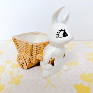 May include: A white ceramic bunny planter with a brown wooden cart. The bunny has black eyelashes and a pink nose. The cart has a rectangular opening for planting. The planter is on a floral patterned surface.
