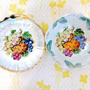 May include: Two vintage decorative plates with iridescent rims and fruit designs. Each plate features a pineapple, grapes, and leaves in shades of orange, purple, green, and blue. One plate has a scalloped gold rim.