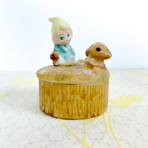 Puede incluir: Una caja de baratijas de cerámica vintage con una figura de gnomo sentado y un perro pequeño encima. La caja es cilíndrica con un diseño texturizado, similar a la paja. El gnomo lleva un sombrero amarillo y ropa azul. La caja está pintada en tonos tierra apagados.