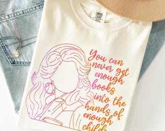 May include: A cream-colored short-sleeve t-shirt featuring a pink and orange gradient line art illustration of a person with long hair and the text: You can never get enough books into the hands of enough children. The shirt has a crew neckline.