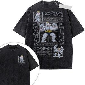 May include: A charcoal grey acid-wash t-shirt featuring a small Machop graphic on the front chest. The back displays large illustrations of Machop, Machoke, and Machamp with descriptive text in English and Japanese, including the names of each character.