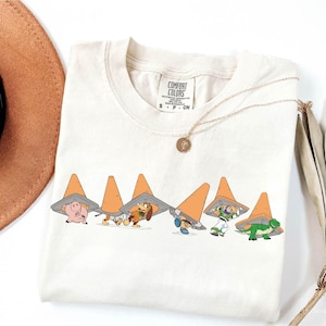 Può includere: Una t-shirt a maniche corte color crema con una stampa grafica di sei personaggi dei cartoni animati, ognuno con un cono stradale arancione sulla testa. La maglietta presenta un'etichetta del marchio Comfort Colors sul collo e una sottile catenina dorata.