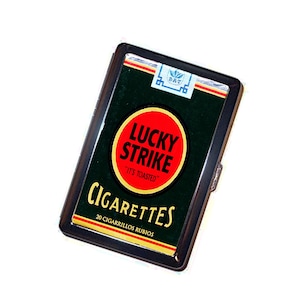 May include: A rectangular metal cigarette case featuring a dark green background with a prominent red circle logo containing the black text LUCKY STRIKE and IT'S TOASTED. Below, the word CIGARETTES appears in gold, with 20 CIGARRILLOS RUBIOS below.
