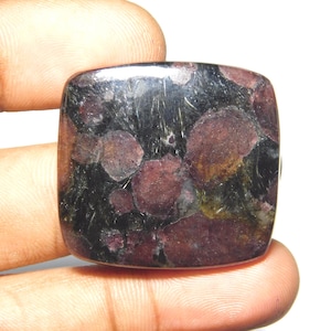 Puede incluir: Una piedra preciosa rectangular pulida con esquinas redondeadas, que presenta una superficie oscura y moteada con inclusiones de color burdeos profundo y negro. La piedra muestra un acabado liso y reflectante con patrones minerales.