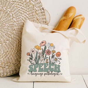 Op de afbeelding: Een witte canvas tote bag met een bloemenontwerp en de tekst "SPEECH language pathologist".