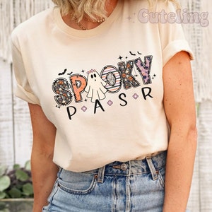 May include: Cream-colored t-shirt with the words "Spooky P A S R" in a Halloween-themed design. The word "Spooky" features a mix of patterns, including pumpkins and bats. A ghost graphic is between the letters "P" and "A".