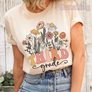 Op de afbeelding: Een beige T-shirt met een bloemenprint en de tekst "THIRD grade" in roze, gele en oranje letters.