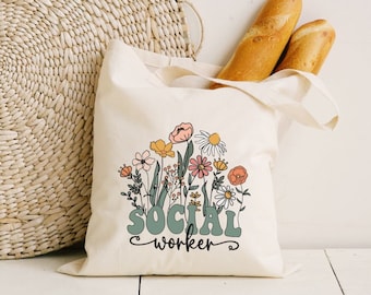 Könnte beinhalten: Eine weiße Canvas-Tragetasche mit einem Blumendesign und dem Text "SOCIAL worker" in grünen Buchstaben. Die Tasche liegt auf einer Holzoberfläche mit zwei Baguettes dahinter.