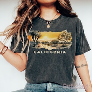 May include: A dark gray t-shirt with a watercolor illustration of a California sunset and the word "CALIFORNIA" printed in yellow. The shirt is worn by a person with a gold necklace and blue jeans.