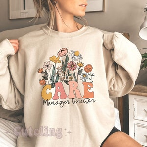 Könnte beinhalten: Ein beiger Pullover mit einem Blumendesign und dem Text "CARE Manager Director".
