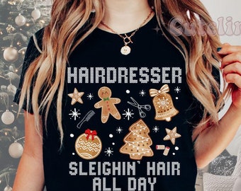 Op de afbeelding: Zwart T-shirt met een feestelijk ontwerp voor kappers. Het shirt heeft de tekst "HAIRDRESSER SLEIGHIN' HAIR ALL DAY" met peperkoek-thema graphics. De zin "MORE COLORS AVAILABLE" wordt ook weergegeven.