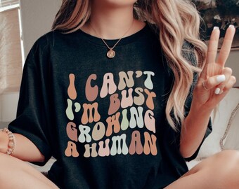 May include: A black t-shirt with a colorful graphic that reads "I can't I'm busy growing a human".