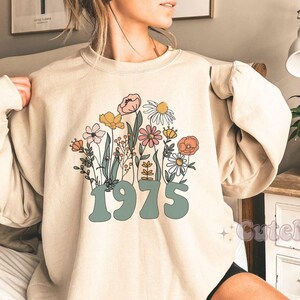 Könnte beinhalten: Ein beiger Pullover mit einem Blumendesign und der Jahreszahl 1975 in Grün gedruckt.