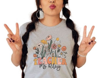 Pode incluir: Uma camiseta cinza com um design floral e o texto "TEACHER Ms Riley" em letras coloridas.