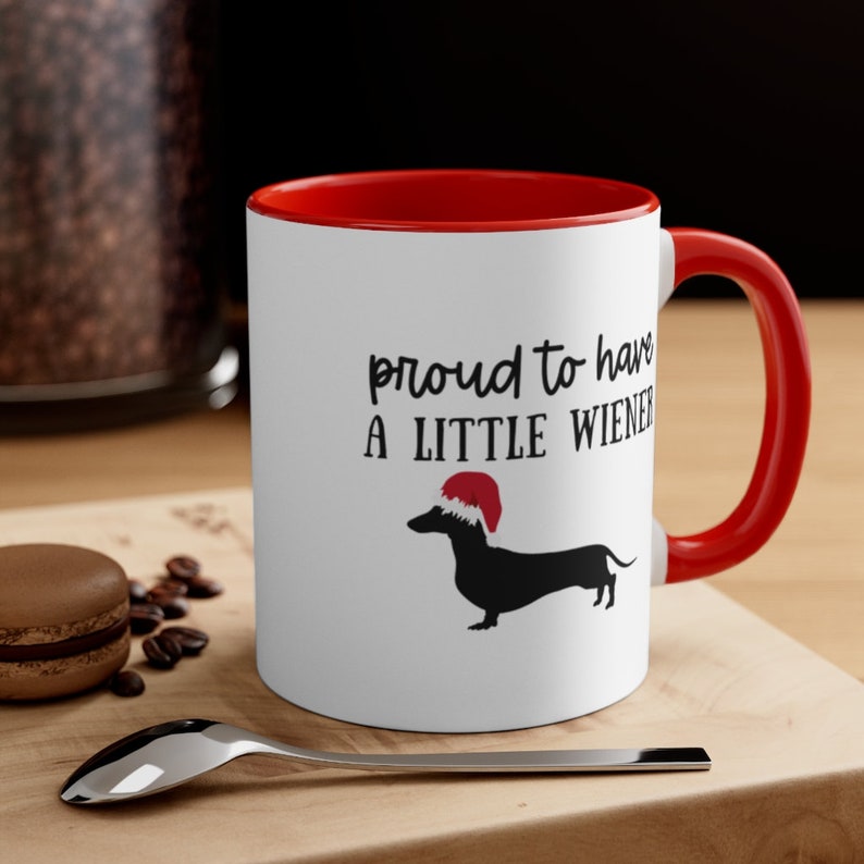 May include: White ceramic mug with a red interior and handle. The mug features the text "proud to have a little wiener" with a black dachshund wearing a Santa hat. The mug is on a wooden surface.