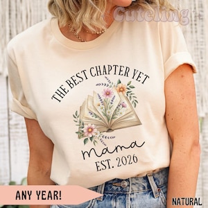 Może przedstawiać: Kremowa koszulka z napisem "THE BEST CHAPTER YET" nad ilustracją otwartej książki ozdobionej kwiatami. Pod książką napis "mama" i "EST. 2026". Na dole koszulki napis "ANY YEAR!" i "NATURAL".