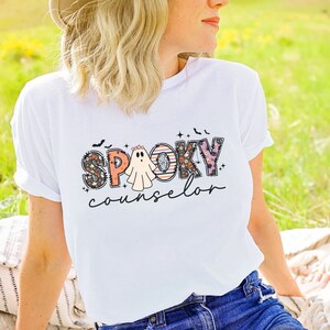 May include: White t-shirt with a graphic design that says "Spooky Counselor" in colorful patchwork letters with a ghost in the middle.