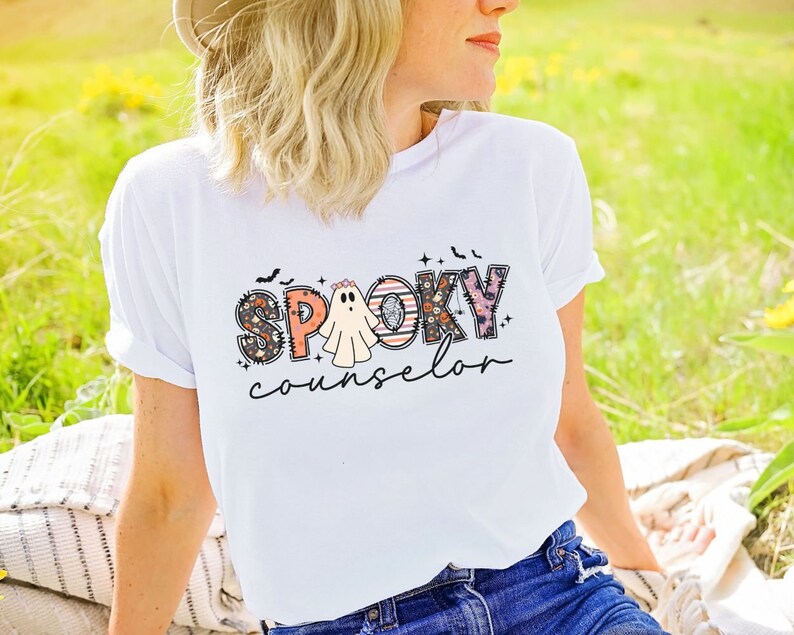 May include: White t-shirt with a graphic design that says "Spooky Counselor" in colorful patchwork letters with a ghost in the middle.