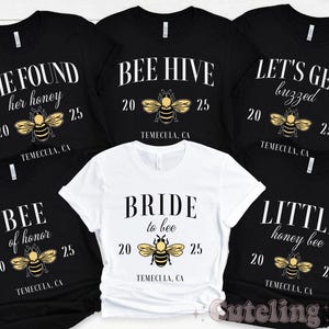 May include: Six black t-shirts with white text and bee graphics. The shirts say "She Found Her Honey", "Bee Hive", "Let's Get Buzzed", "Bee of Honor", "Bride to Bee", and "Little Honey Bee".