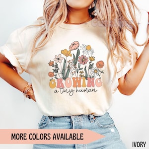 Könnte beinhalten: Elfenbeinfarbenes T-Shirt mit floralem Design und dem Text "GROWING a tiny human" in Retro-Schrift. Das Design zeigt rosa, gelbe und orangefarbene Blumen. Ein Banner unten zeigt "MORE COLORS AVAILABLE".