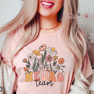 Puede incluir: Una camiseta rosa con un dise&ntilde;o floral y el texto "NEURO team" en letras de colores.