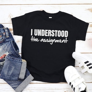 Könnte beinhalten: Schwarzes T-Shirt mit weißem Text, der "Ich habe die Aufgabe verstanden" lautet. Das Shirt liegt auf einer weißen Holzoberfläche mit einer blauen Jeans und weißen Turnschuhen.