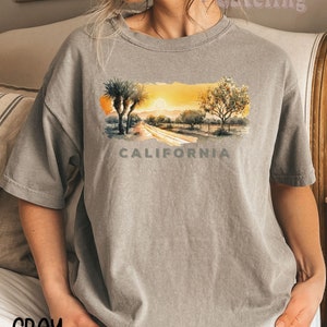 May include: Gray t-shirt with a watercolor illustration of a California landscape with palm trees, a road, and the word "CALIFORNIA" printed below the image.