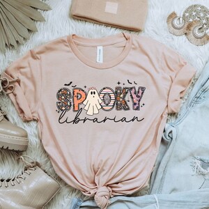 May include: A pink t-shirt with the text "Spooky Librarian" in colorful letters with a ghost in the middle of the word "Spooky".