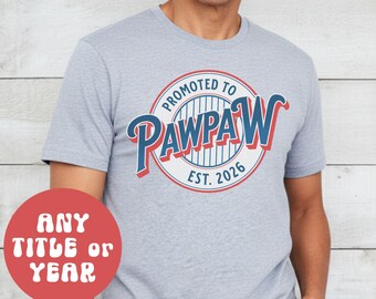 Puede incluir: Camiseta gris deportiva con un gráfico circular que dice "Promoted to Pawpaw Est. 2026" en rojo y azul. Un círculo rojo con texto blanco que dice "Any Title or Year" está en la esquina inferior izquierda.