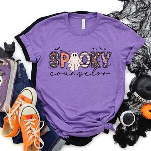 May include: Purple t-shirt with a black and white ghost and the text "Spooky Counselor" in colorful patchwork letters.