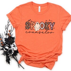 May include: Orange t-shirt with a graphic design of the word "spooky" in colorful letters with a ghost in the middle and the word "counselor" below.