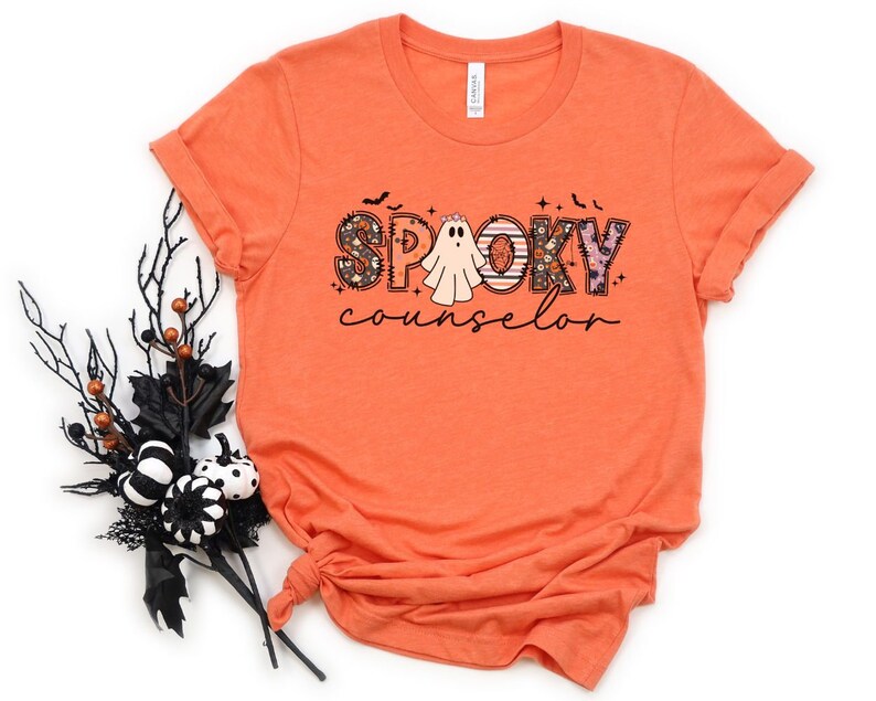 May include: Orange t-shirt with a graphic design of the word "spooky" in colorful letters with a ghost in the middle and the word "counselor" below.