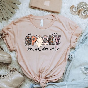May include: A light pink t-shirt with the text "Spooky mama" in colorful letters with a ghost in the middle of the word "Spooky".