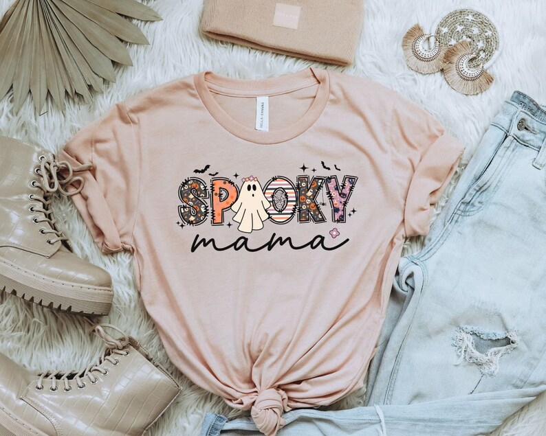 May include: A light pink t-shirt with the text "Spooky mama" in colorful letters with a ghost in the middle of the word "Spooky".