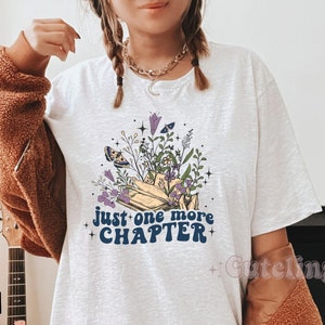 Puede incluir: Camiseta blanca con un gr&aacute;fico de un libro con flores y mariposas. El texto "just one more chapter" est&aacute; escrito en letras azules.