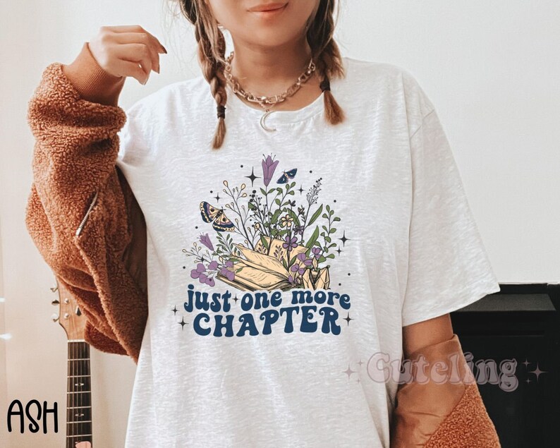 Puede incluir: Camiseta blanca con un gr&aacute;fico de un libro con flores y mariposas. El texto "just one more chapter" est&aacute; escrito en letras azules.