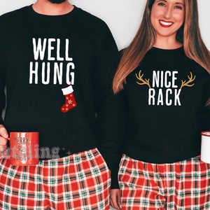 Op de afbeelding: Bijpassende zwarte sweatshirts met witte tekst: "WELL HUNG" en "NICE RACK". Het "WELL HUNG" sweatshirt heeft een rood-witte kerstsok-afbeelding. Beide personen dragen een rood-zwarte geruite pyjamabroek.